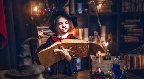 Surprised caucasian little girl in witch Cosplay reads magic book while holding magic wand, Halloween costume. Halloween party.