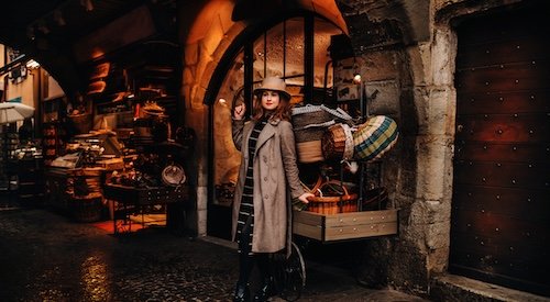 a girl in a coat and hat stands near baskets in the old town of Annecy, spending time outdoors exploring the European city. An elegant young lady in a coat and hat enjoys a view of the old town in France.