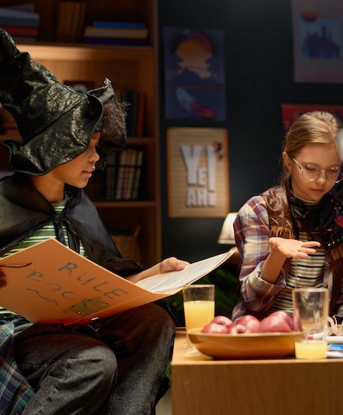 Cute African American boy in witch hat and mantle sitting next to youthful girl during board game and explaining rules from big book